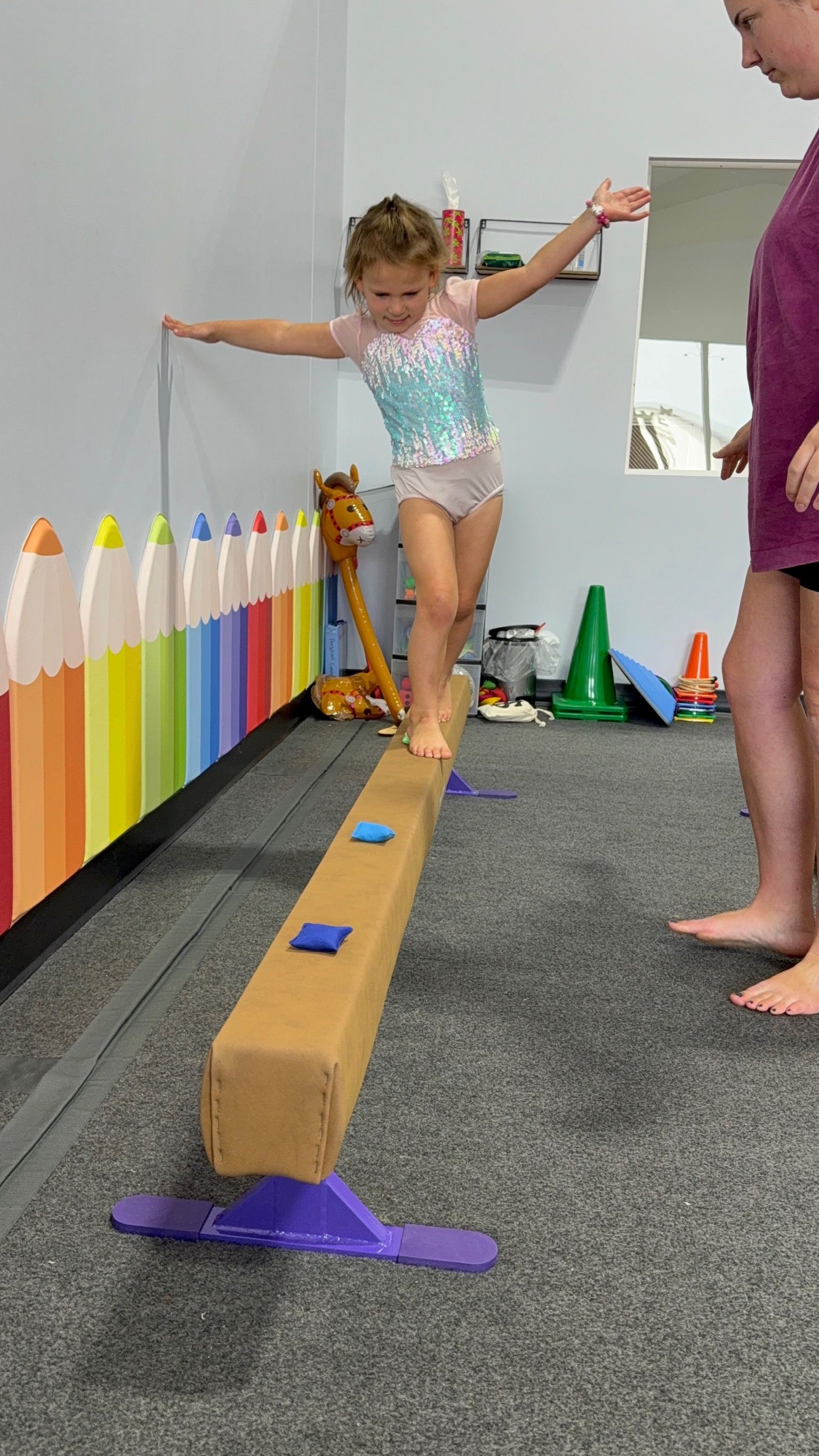 Girl balancing on a beam with arms out for balance, a coach nearby. Gym setting with colorful wall decor.
