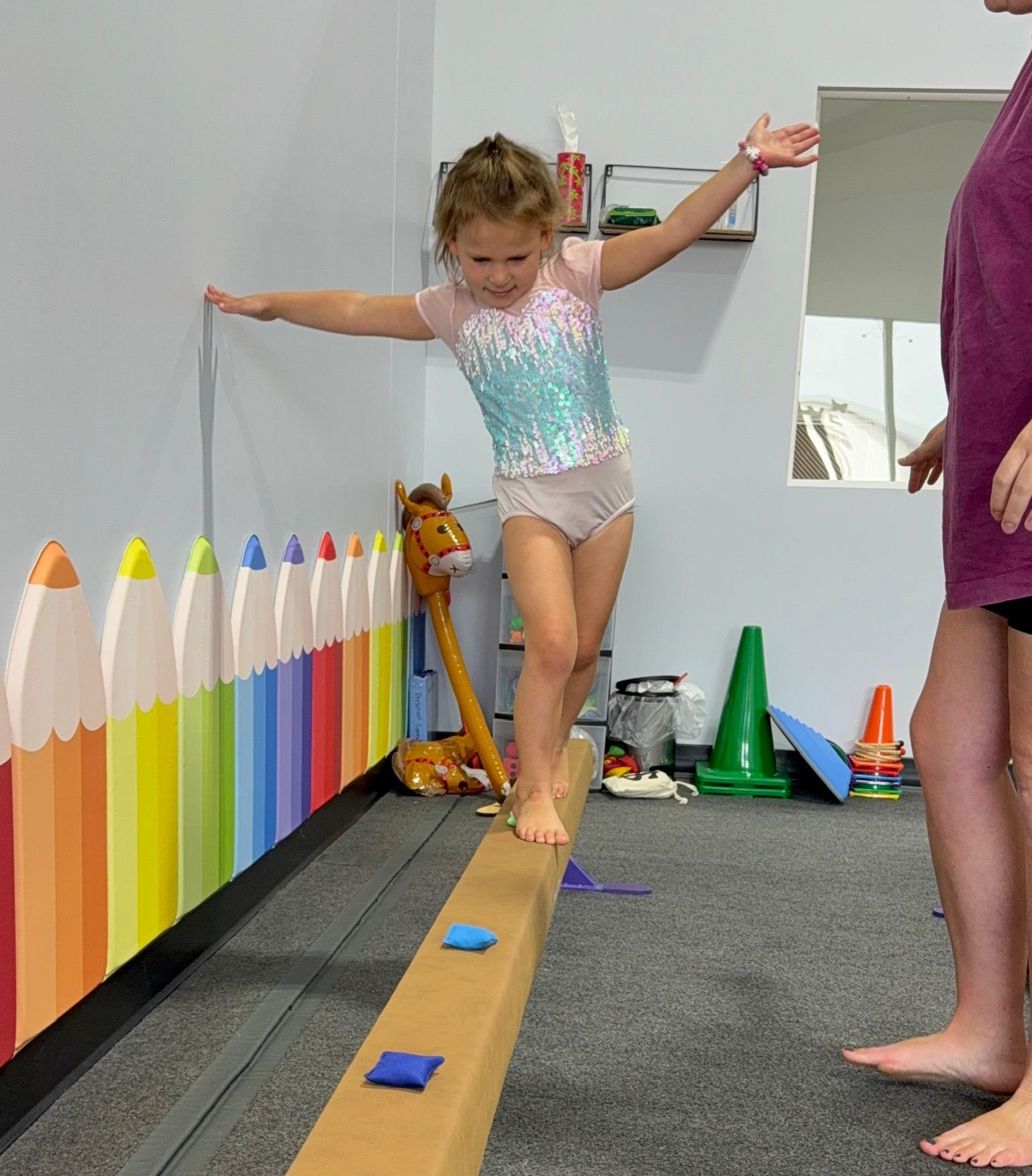 Girl balances on a beam, arms out, in a gym. A person stands nearby. Colorful pencil wall art.