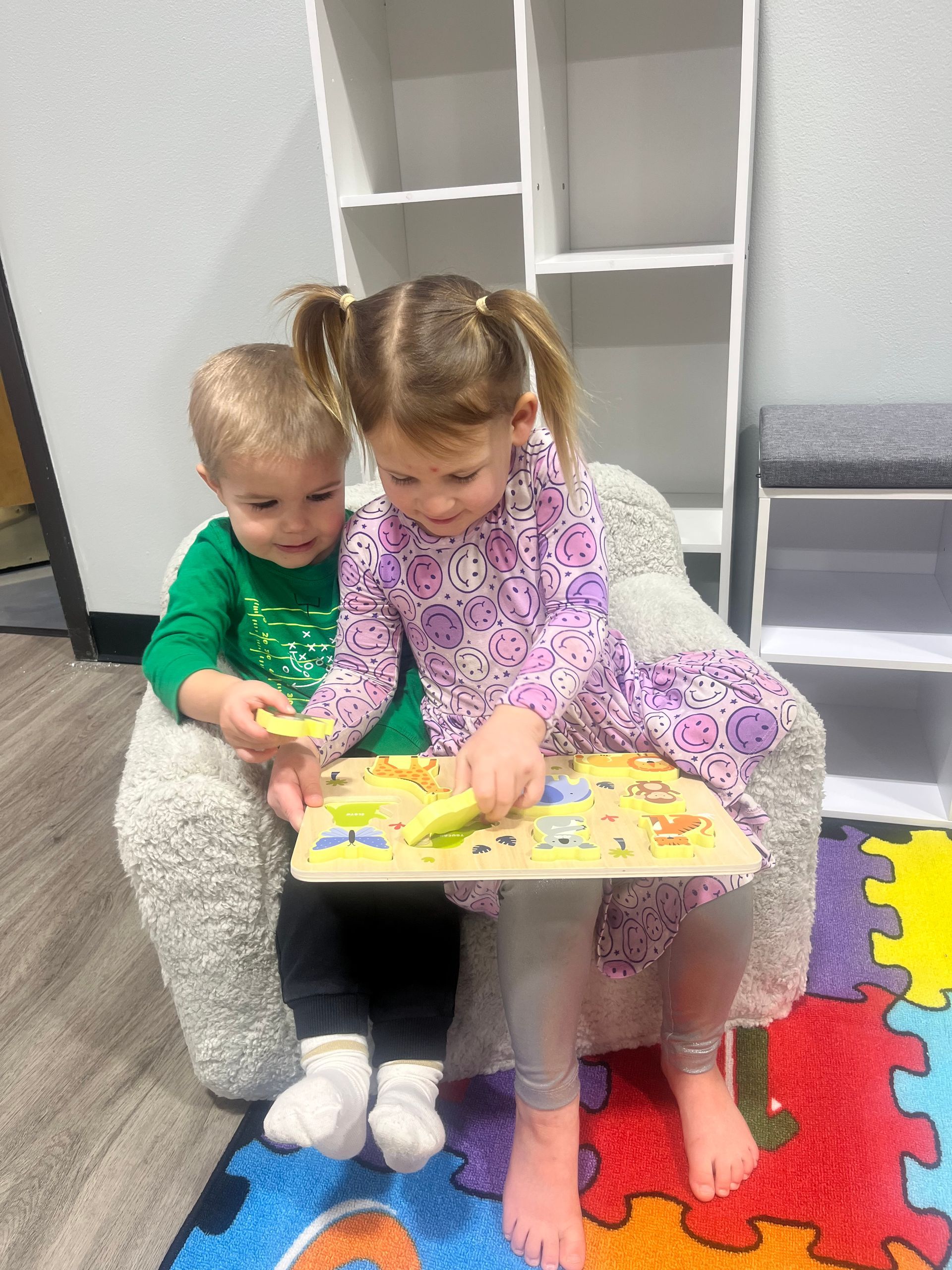 Two children sitting, playing a puzzle. Girl in purple, boy in green. Inside on a fluffy chair.