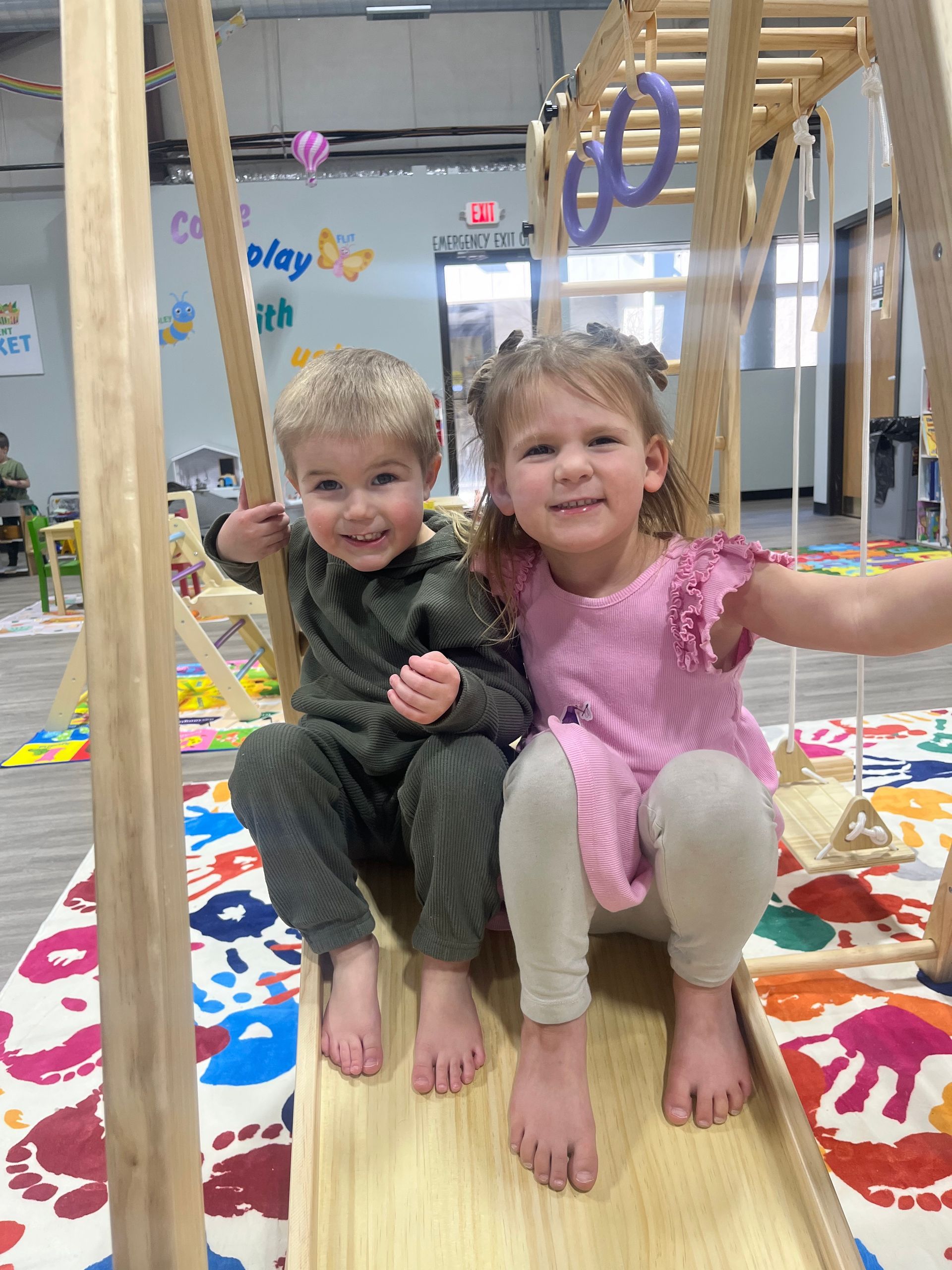 Two smiling children sit on wooden play structure; one in green, one in pink. Brightly patterned rug.