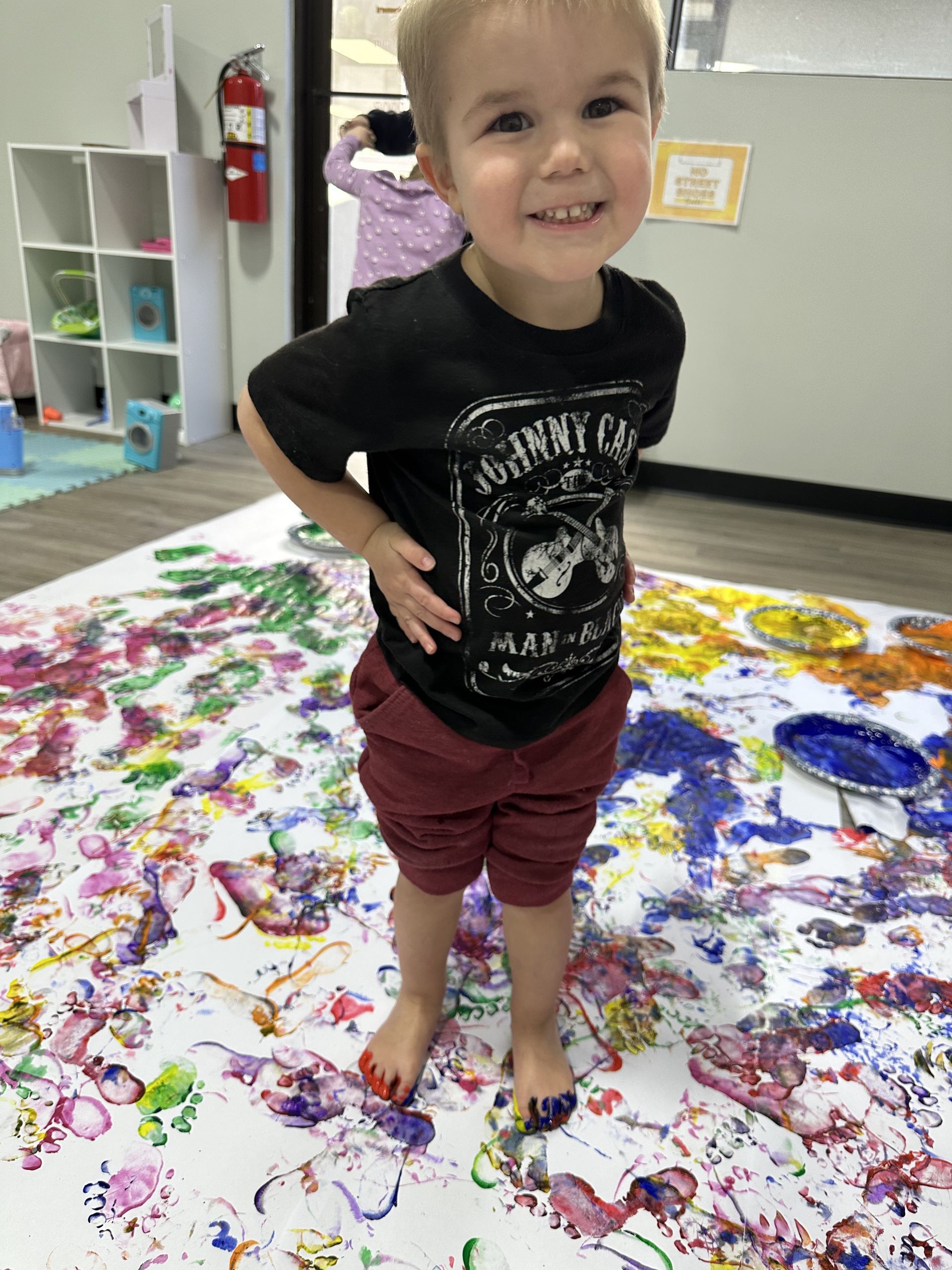 Boy stands on paint-splattered paper, smiling, with paint on his feet. In a playroom.