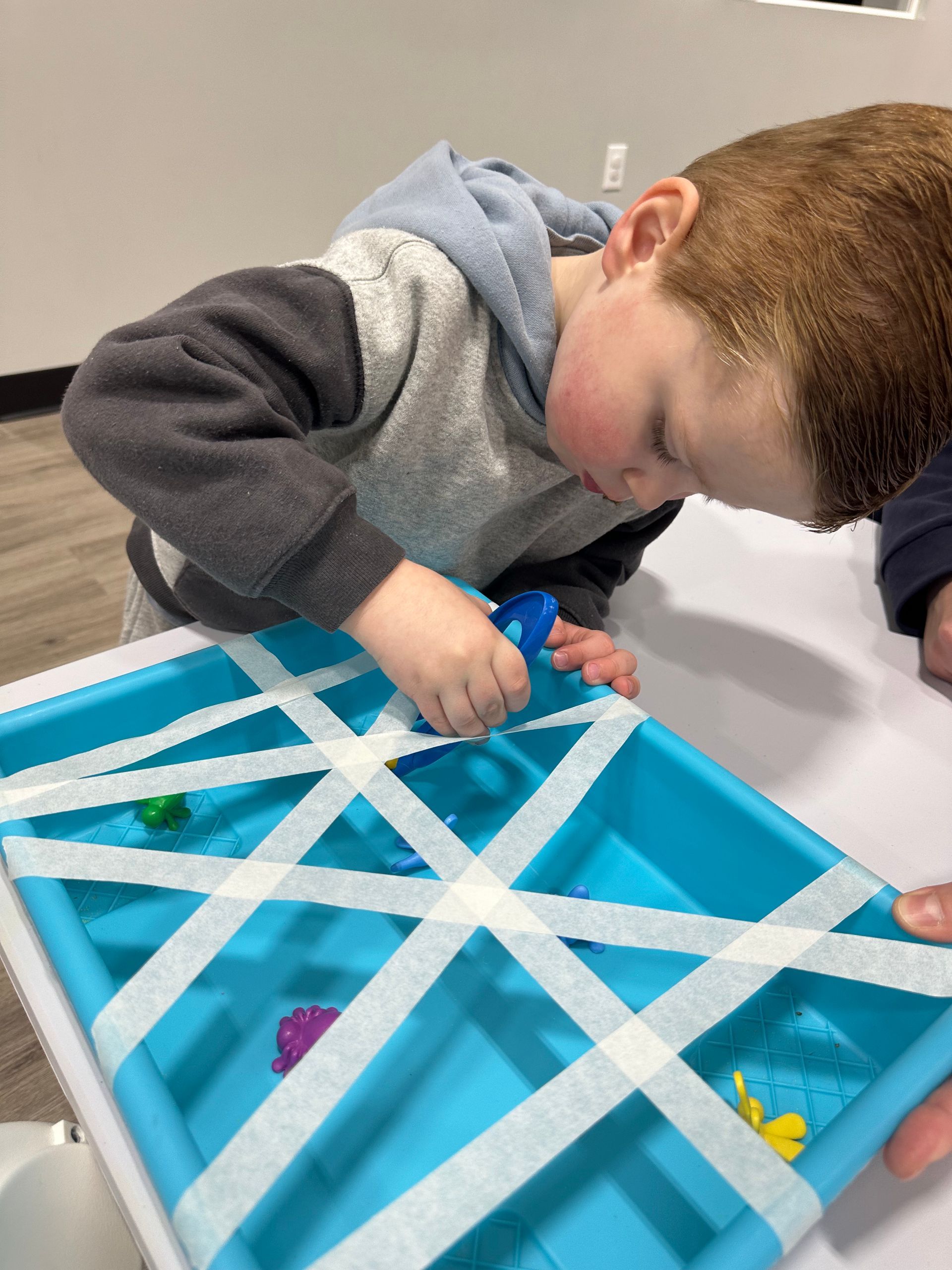 Boy playing with toys in a blue tray. White tape forms sections.