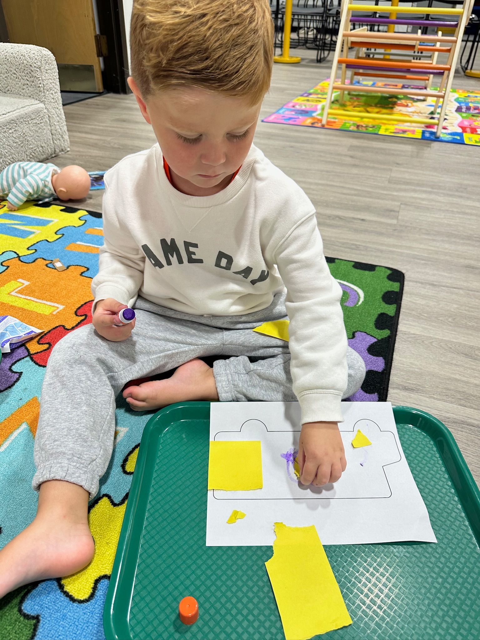 Boy seated on a colorful mat, crafting with paper and glue on a tray.