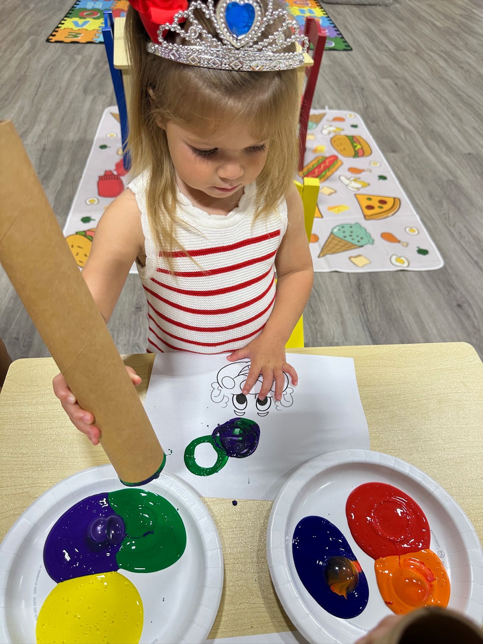 Young child with a tiara making artwork with paint and a cardboard tube.