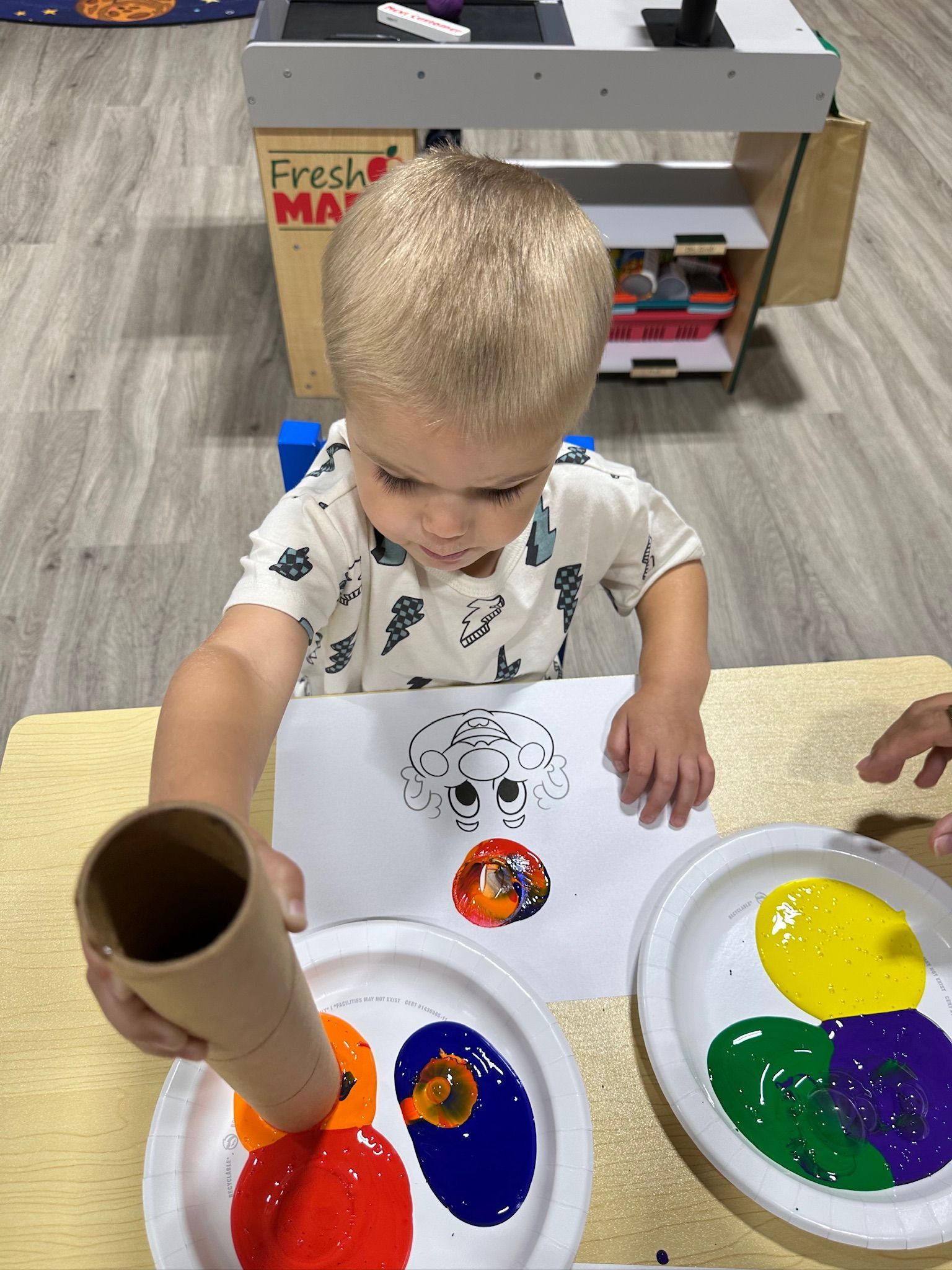 Young child using a cardboard tube to stamp paint onto paper. Paints are red, orange, blue, yellow, and green.