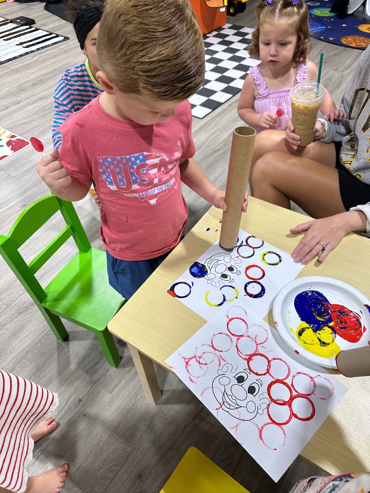 Children doing art project with paint and tube, sitting at a small table.