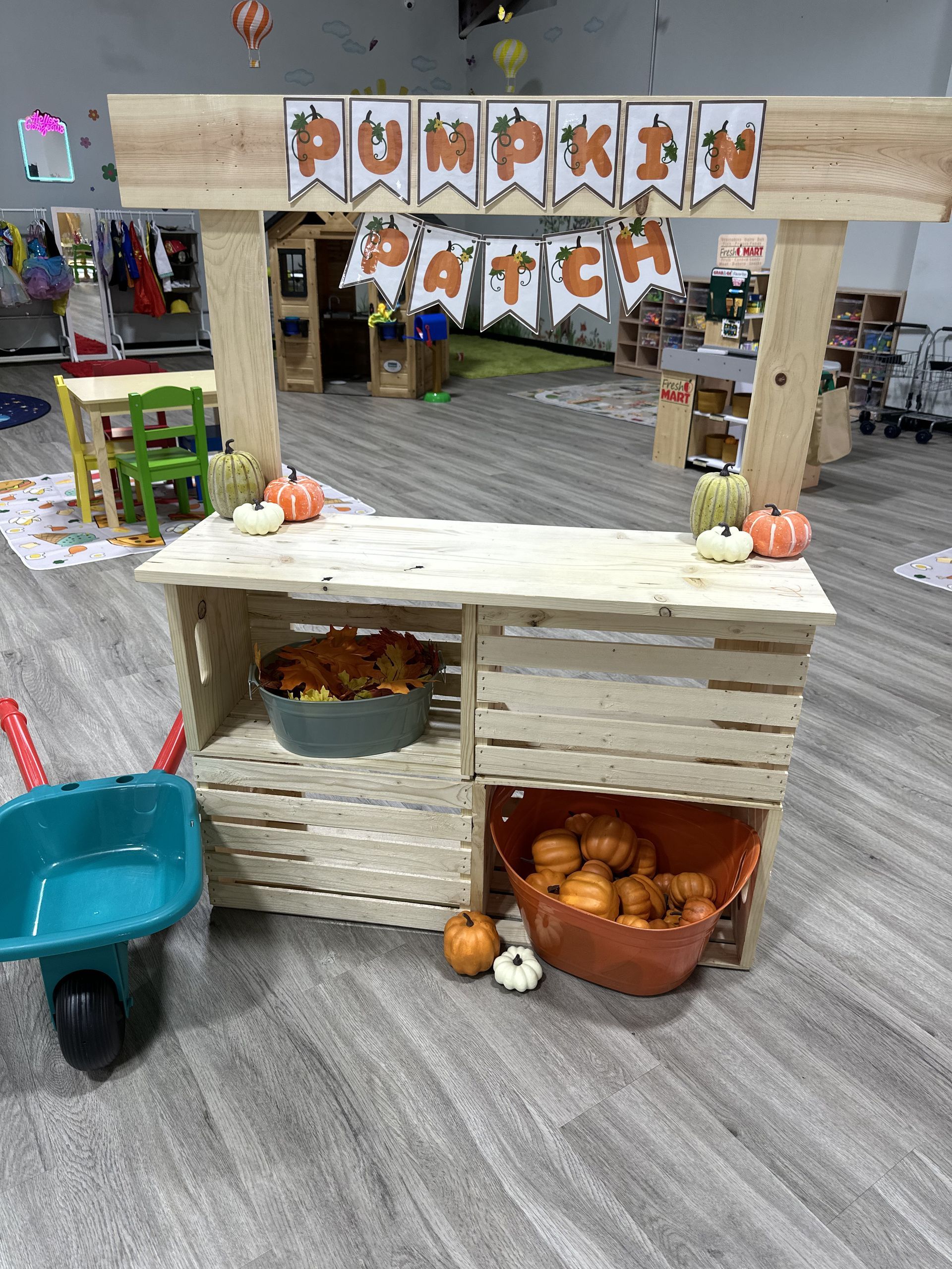Pumpkin patch stand decorated with pumpkins, wooden crates, and a banner. Wheelbarrow nearby.