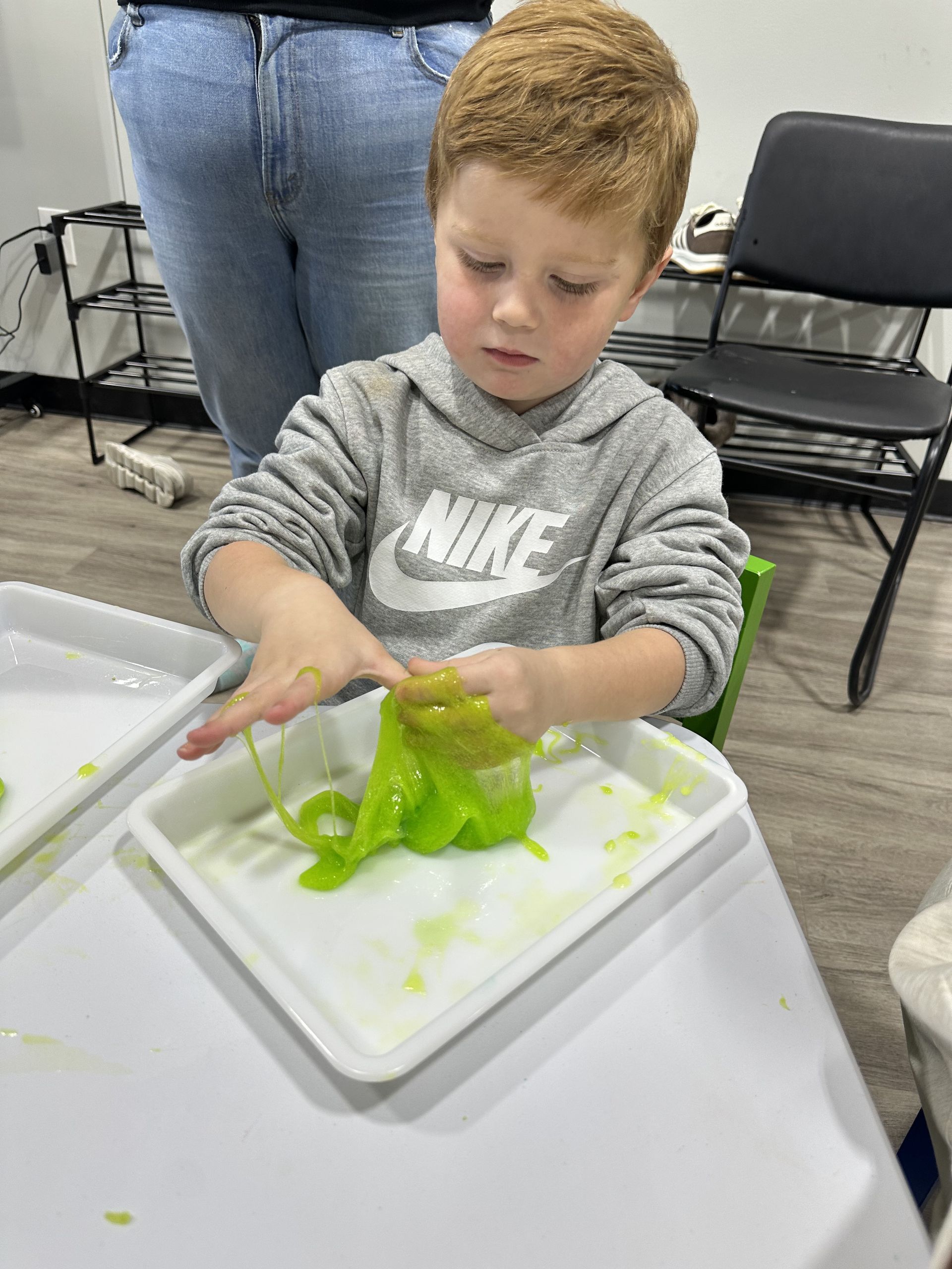 Boy in gray hoodie playing with green slime in a tray at a table.