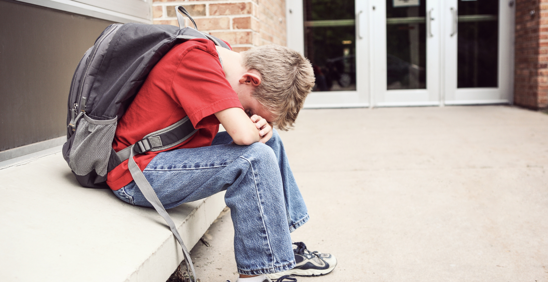 Een jonge jongen met een rugzak zit op een bankje met zijn hoofd naar beneden.