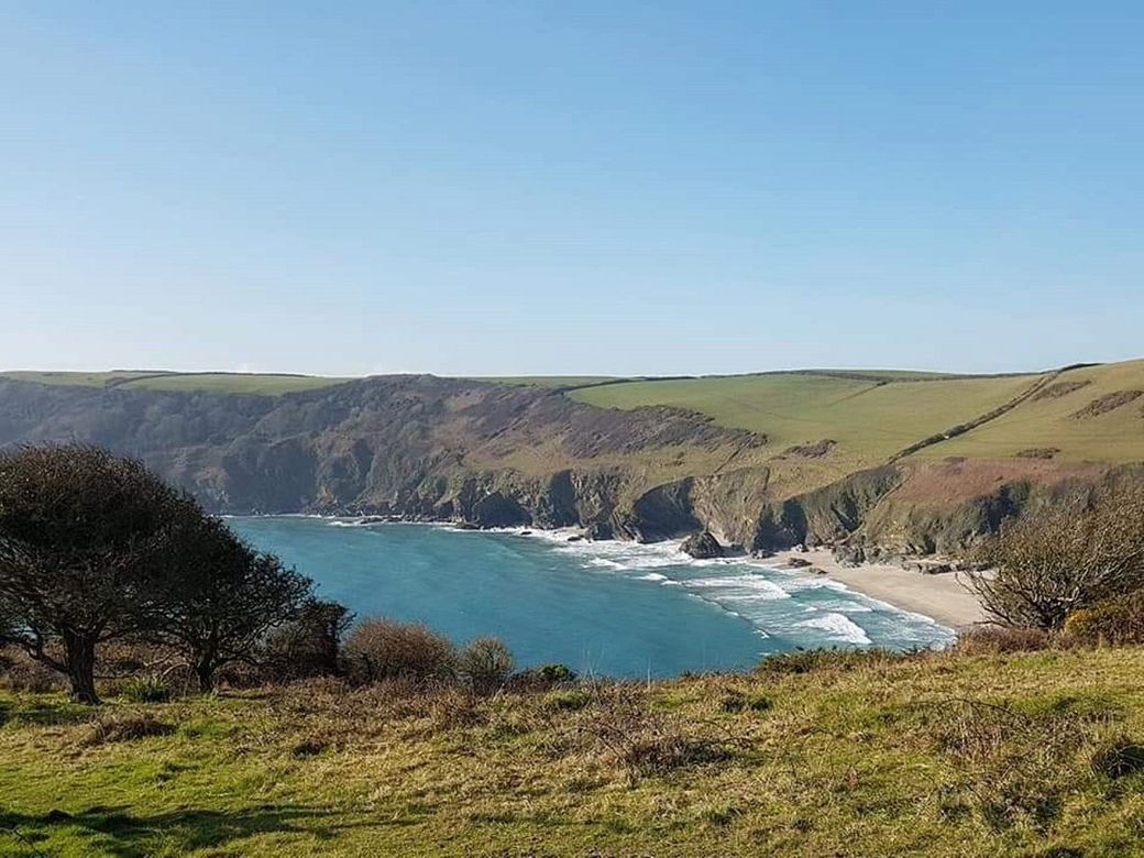 View from a clifftop path into a wide bay with blue water and waves rolling onto a sandy beach