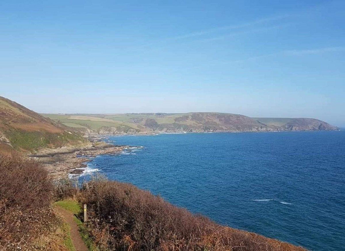 View from a clifftop path into a wide bay with blue water, blue sky and high cliffs beyond