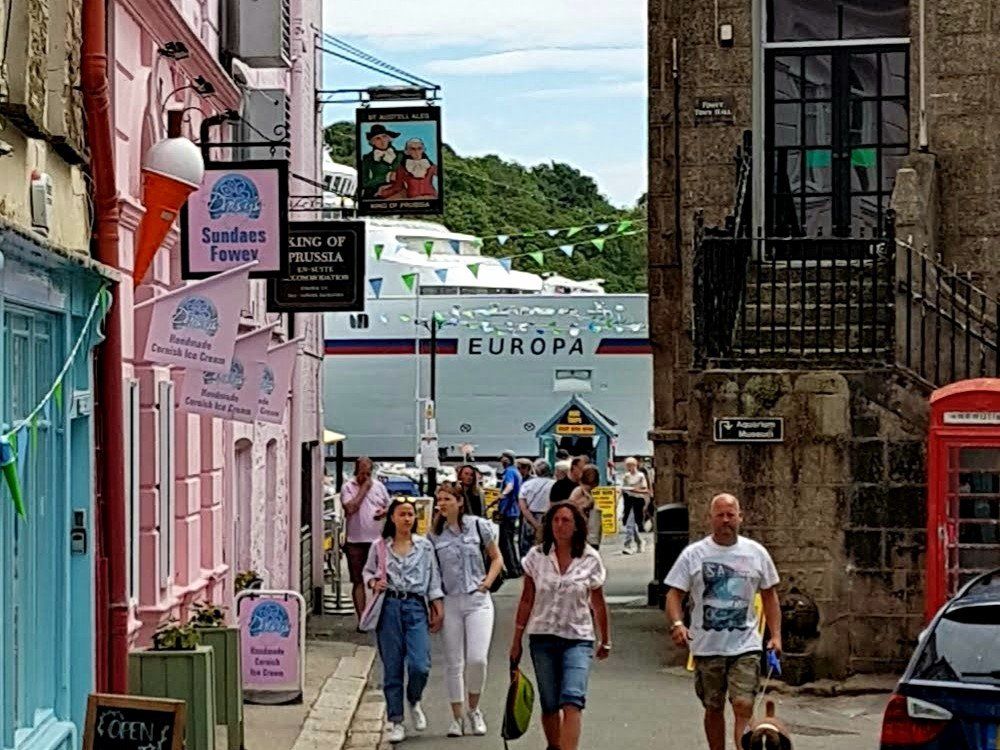 A busy street between ancient buildings looking towards a large cruiseship moored in the river