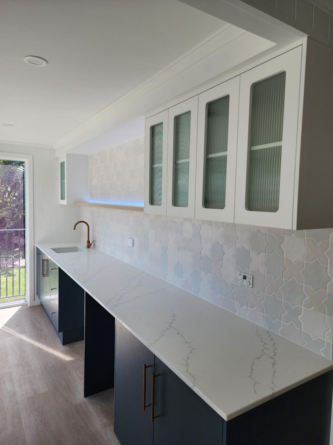 Modern kitchen with long white countertop, dark cabinets, and glass-front upper cupboards by a bright window