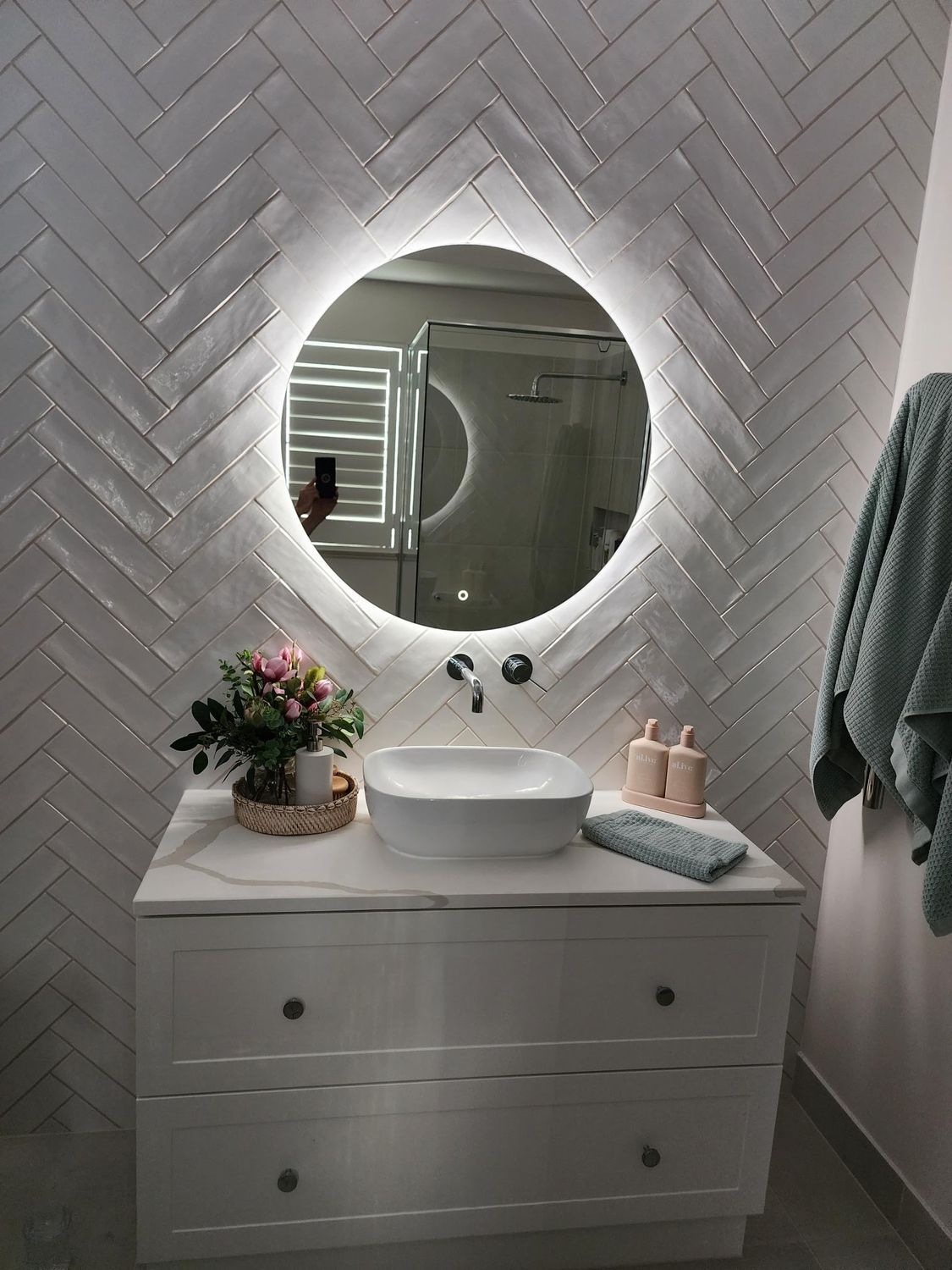Modern bathroom vanity with round mirror, white herringbone tiles, and sink with flowers and soap dispenser
