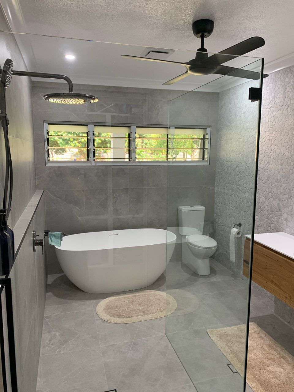 Modern bathroom with freestanding tub, toilet, glass shower, gray walls, wood vanity, and ceiling fan.