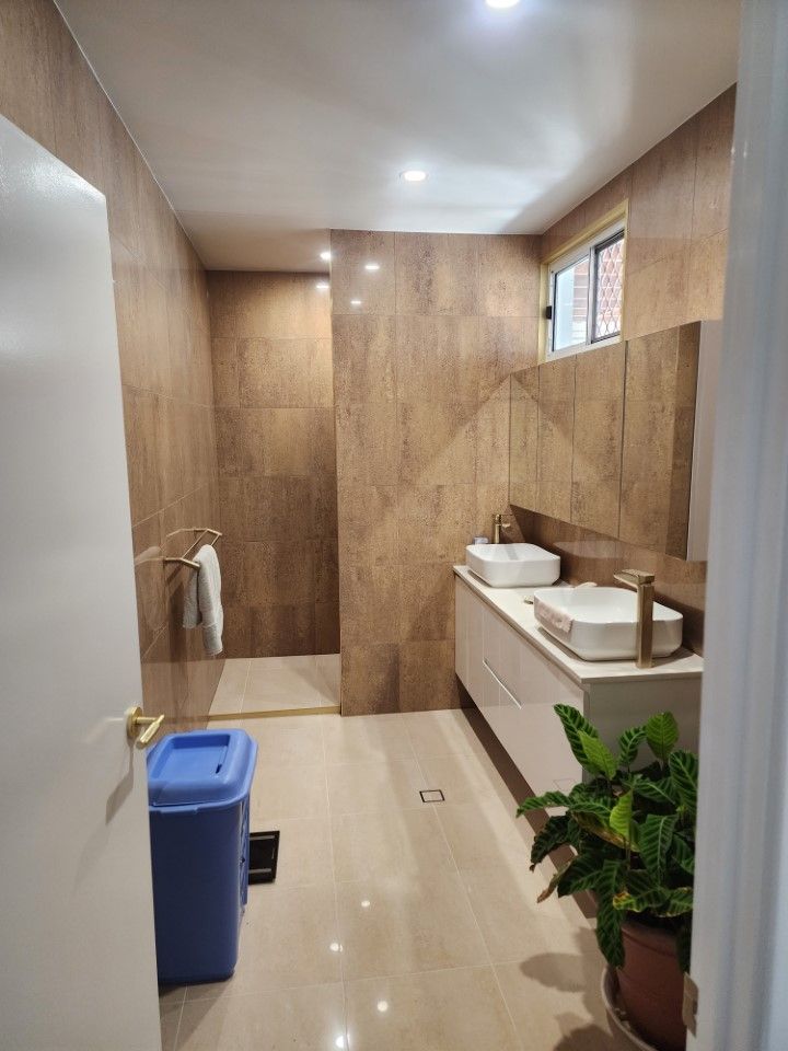 Small restroom with wood-paneled walls, sink counter, blue trash bin, potted plant, and window.