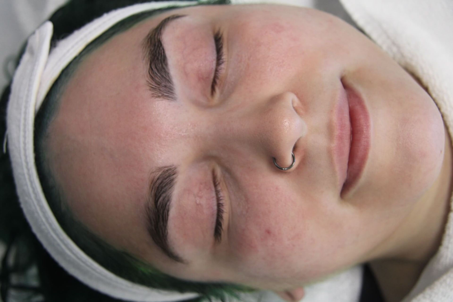 Woman with closed eyes, wearing a white headband, smiles. Freshly done eyebrows are dark.