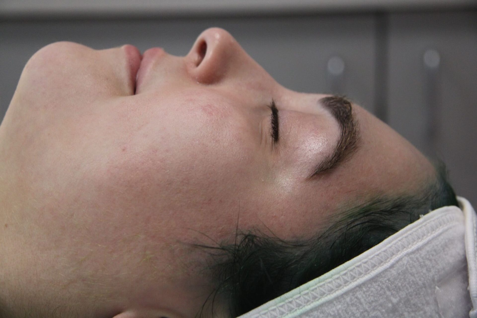 Woman lying down with eyes closed, in a spa-like setting. Light skin, dark eyebrows, with a towel around her head.