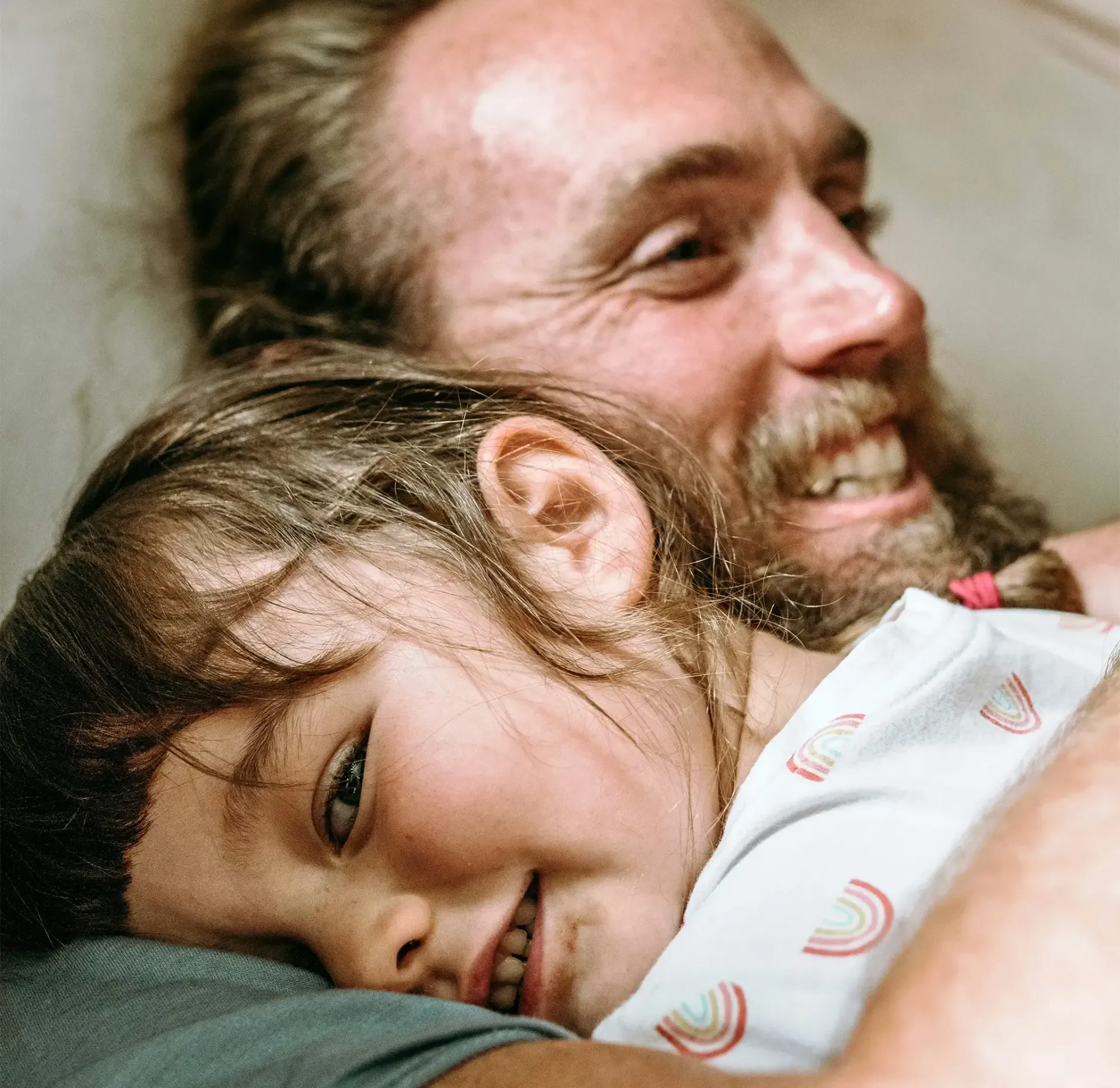Smiling person with a beard hugging a young person with dark hair. Both are smiling, close up.