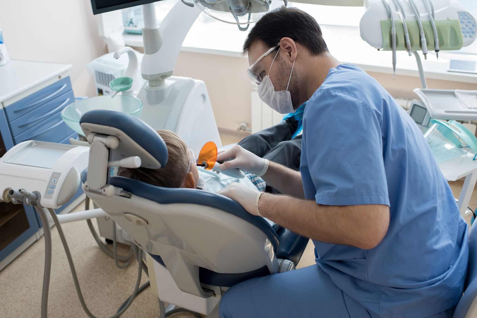 Dentist in blue scrubs working on a patient in a dental chair; bright office setting.