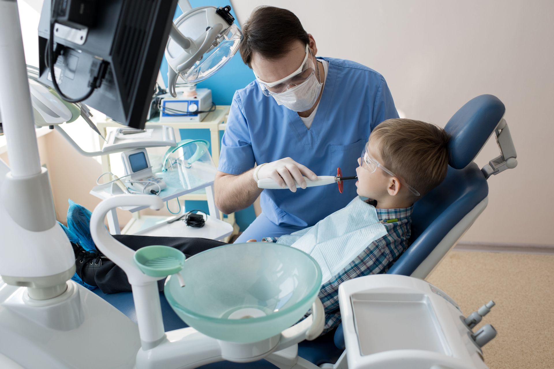 Dentist in blue scrubs examines a child's teeth with a dental tool in a dental office.