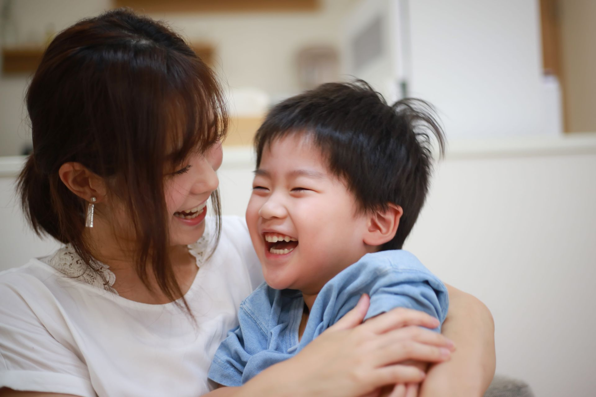 Woman hugging and laughing with a smiling child indoors; neutral background.