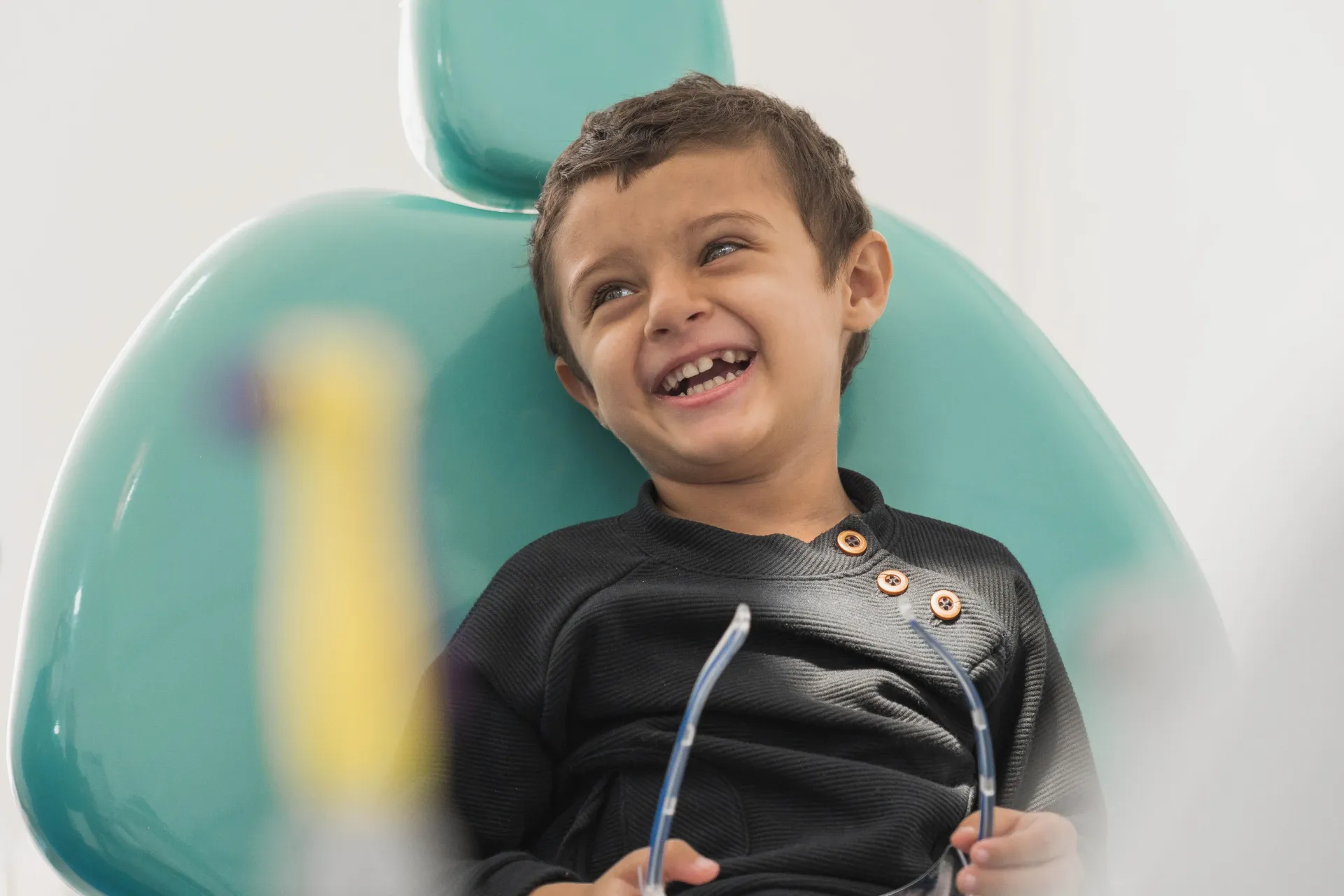Boy laughing, sitting in a dentist's chair, holding glasses. Turquoise chair, black sweater.