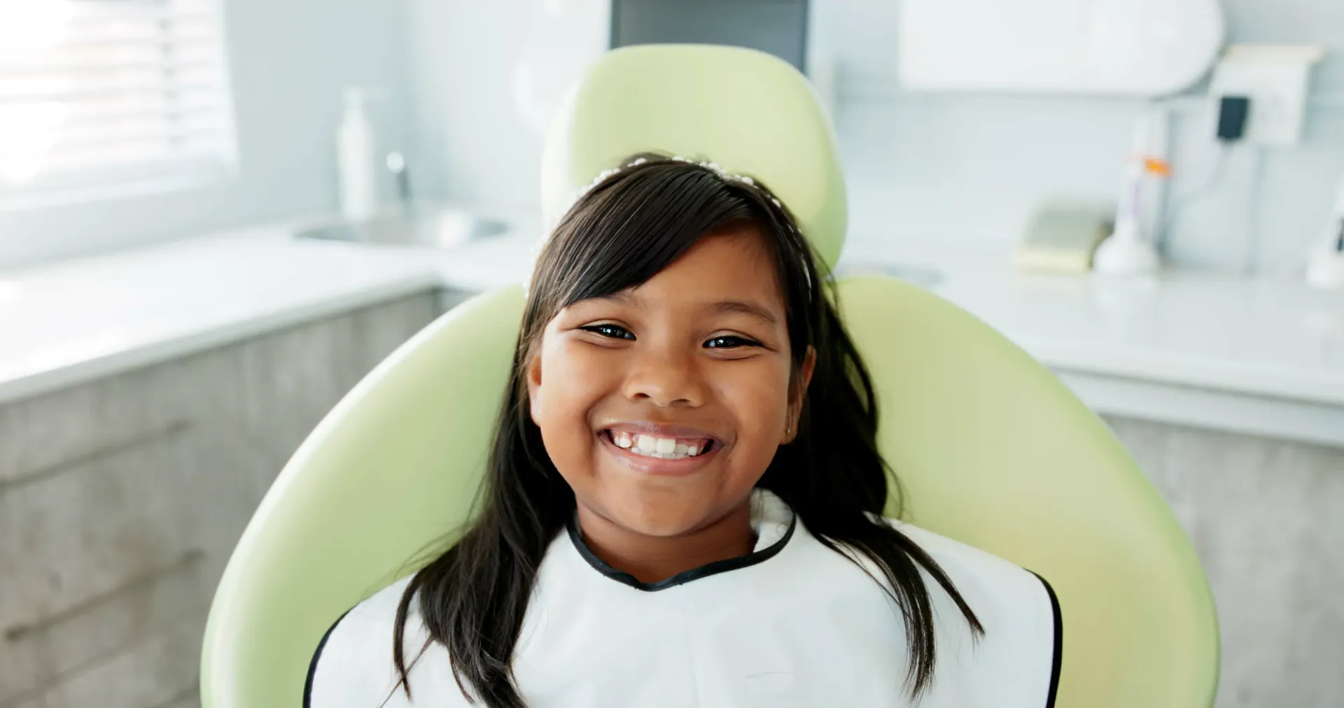 Smiling child in a dental chair wearing a protective bib. Brightly lit room with dental equipment.