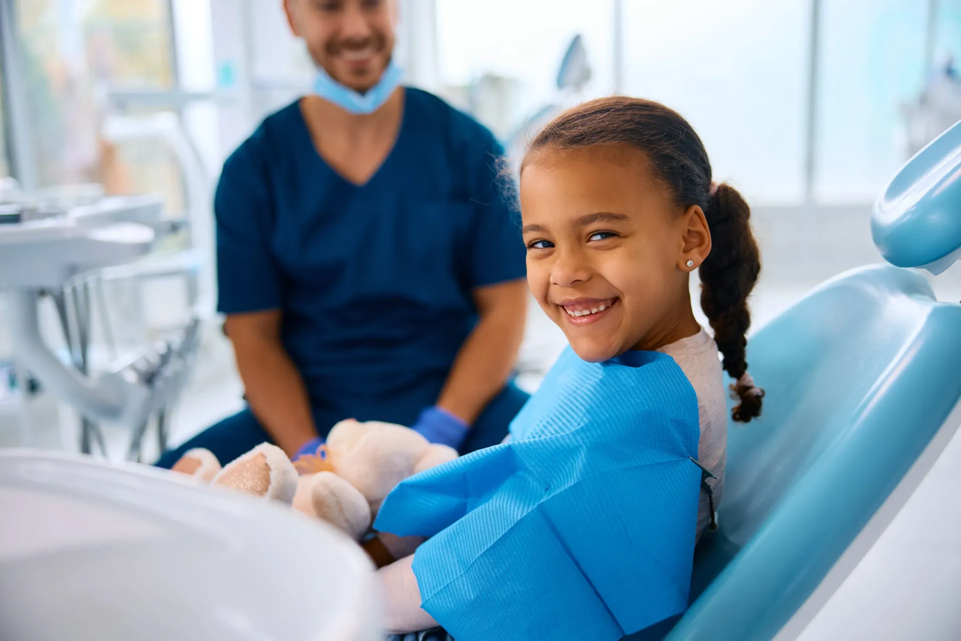 Girl smiling in a dental chair; dentist in background.
