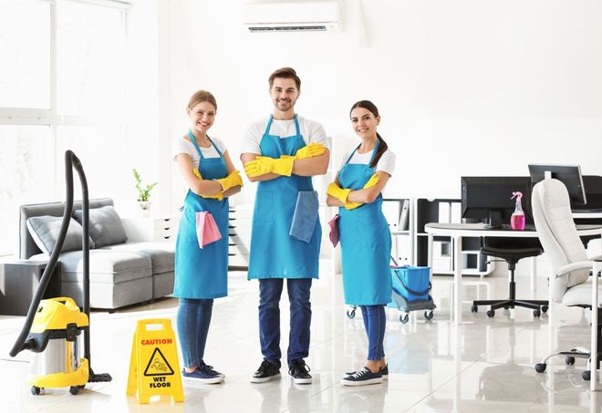 Three smiling cleaners pose in an office. They wear blue aprons, gloves, and stand by a vacuum and wet floor sign.