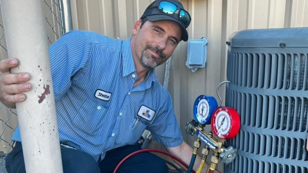 HVAC technician in blue shirt checks AC unit with gauges. He smiles, holding a pipe, outdoors.