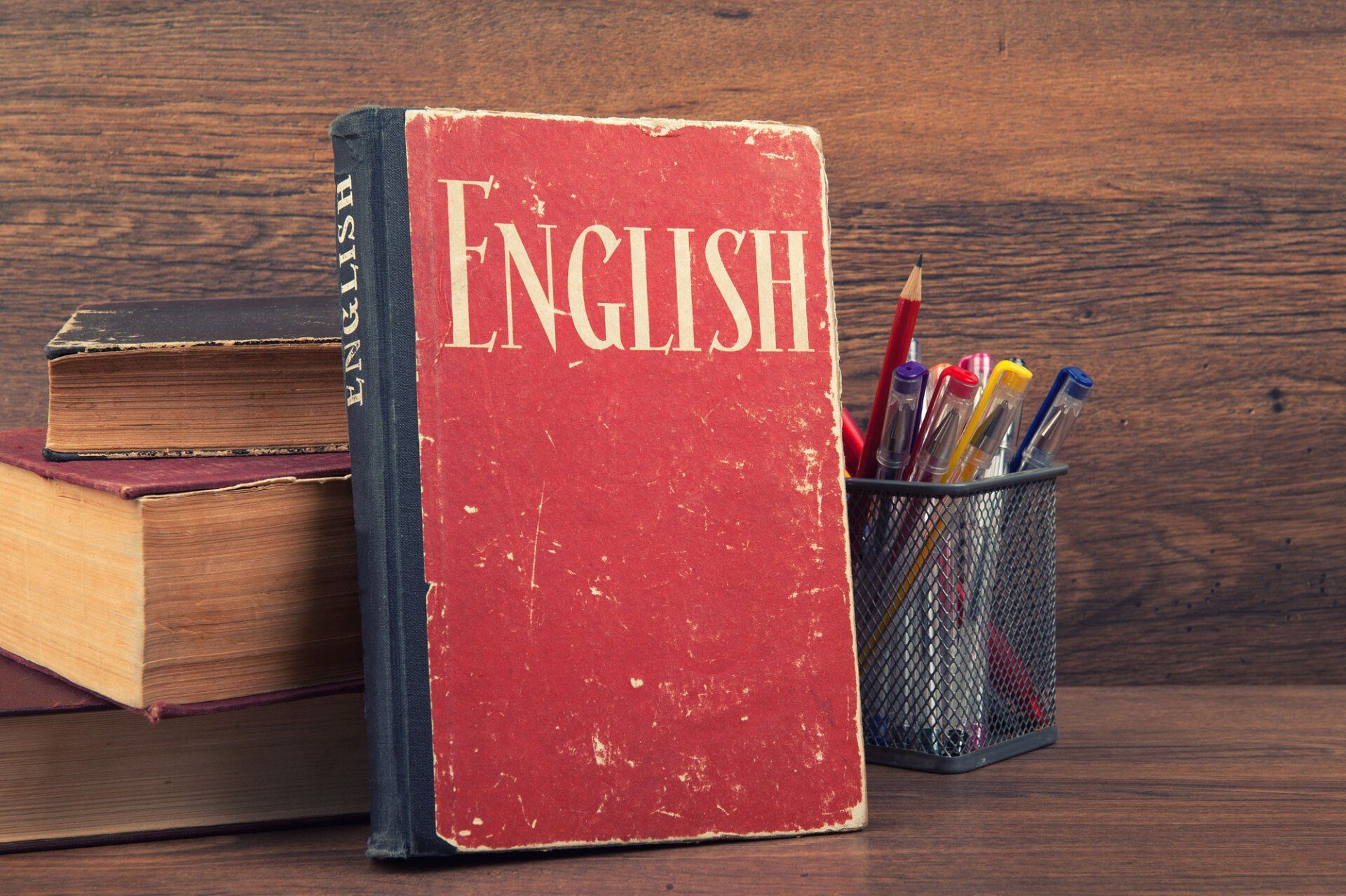 Red English textbook with other books and pens on a wooden surface.