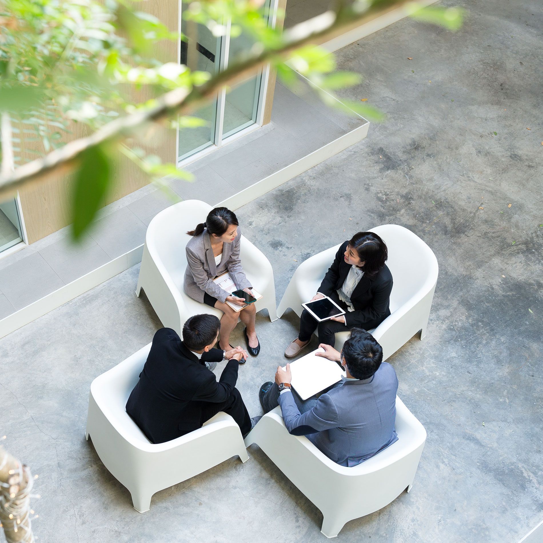 Four people in business attire seated in white chairs, conversing in an outdoor atrium.