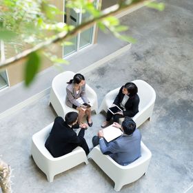 Four people in business attire seated in white chairs, conversing in an outdoor atrium.