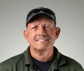 Man with cap and sunglasses smiles, wearing a green shirt, set against a grey background.