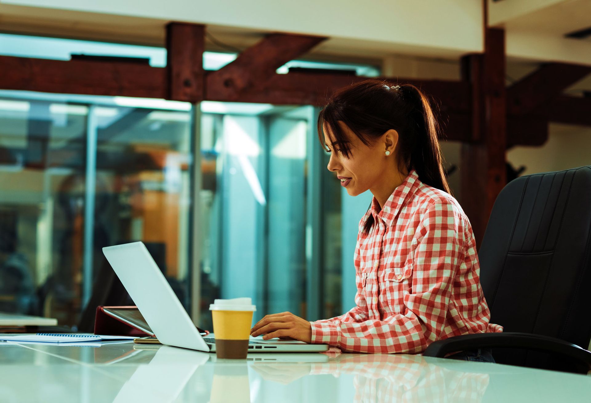 Woman in a checkered shirt working on a laptop at a desk with a coffee cup in a modern office.