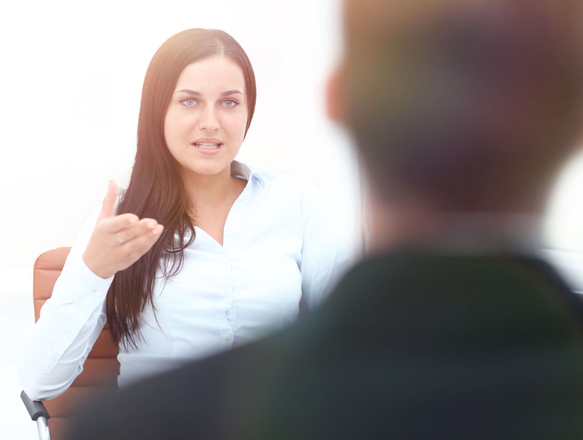 Woman in white shirt gesturing, speaking to person out of focus, seated in an office.