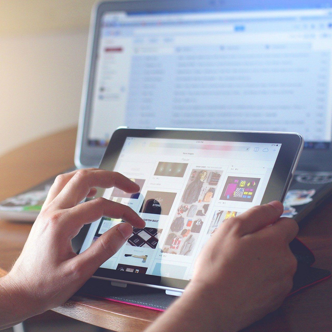 Hands using tablet with website displayed, laptop in background.