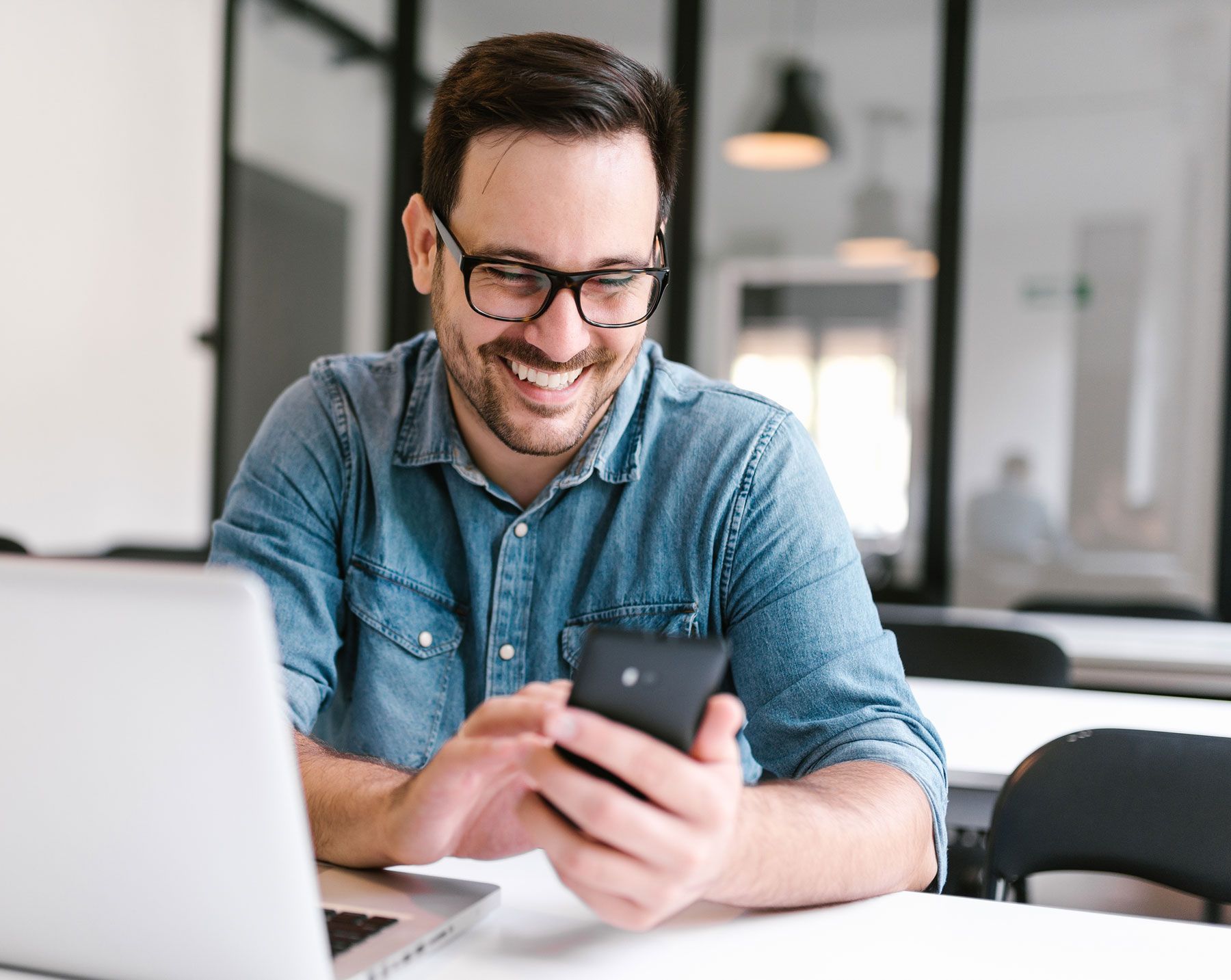 Man with glasses smiling while looking at a phone, laptop open in front of him.