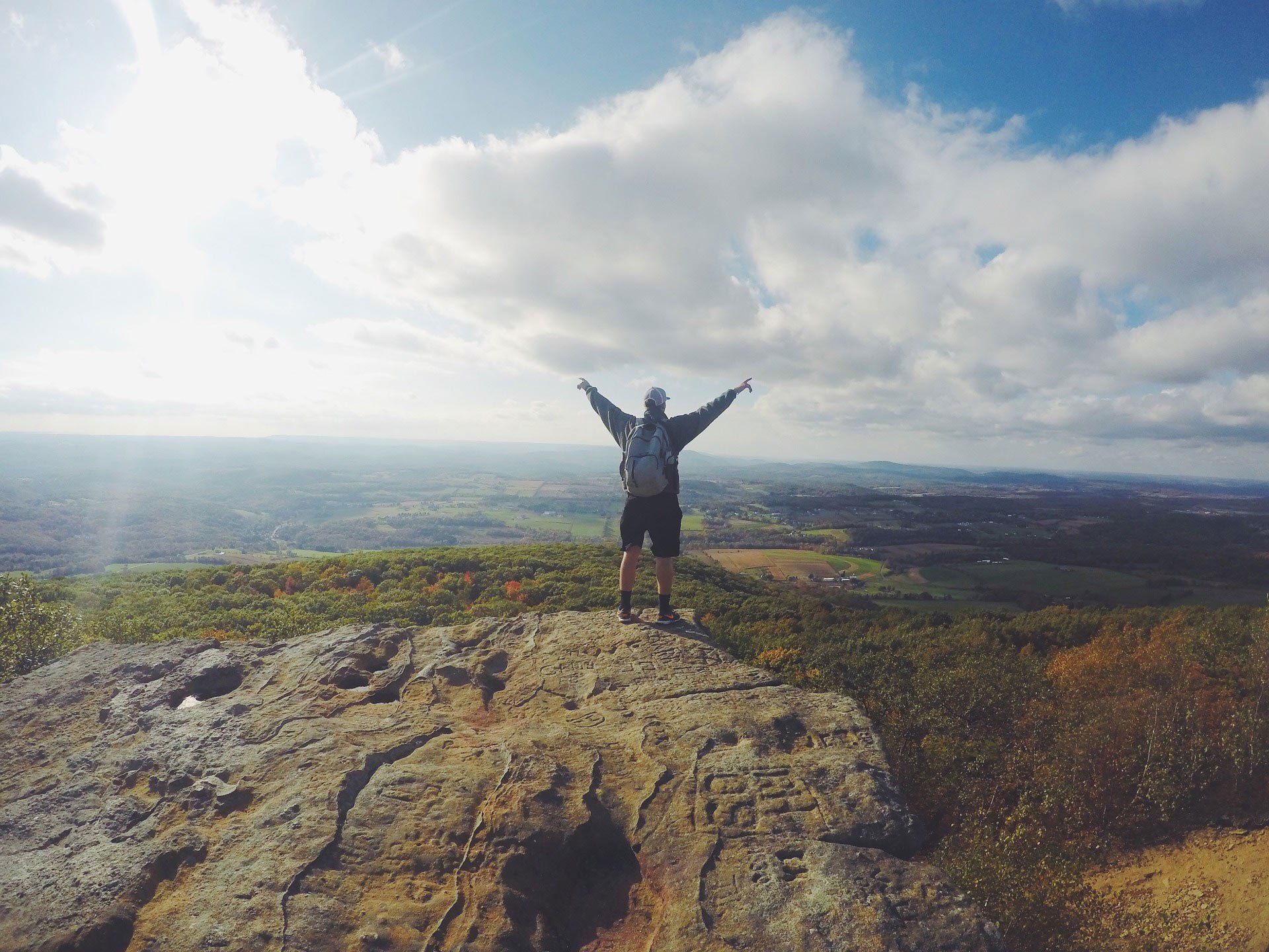 Person with arms raised on a rocky mountaintop, overlooking a scenic valley under a bright, cloudy sky.