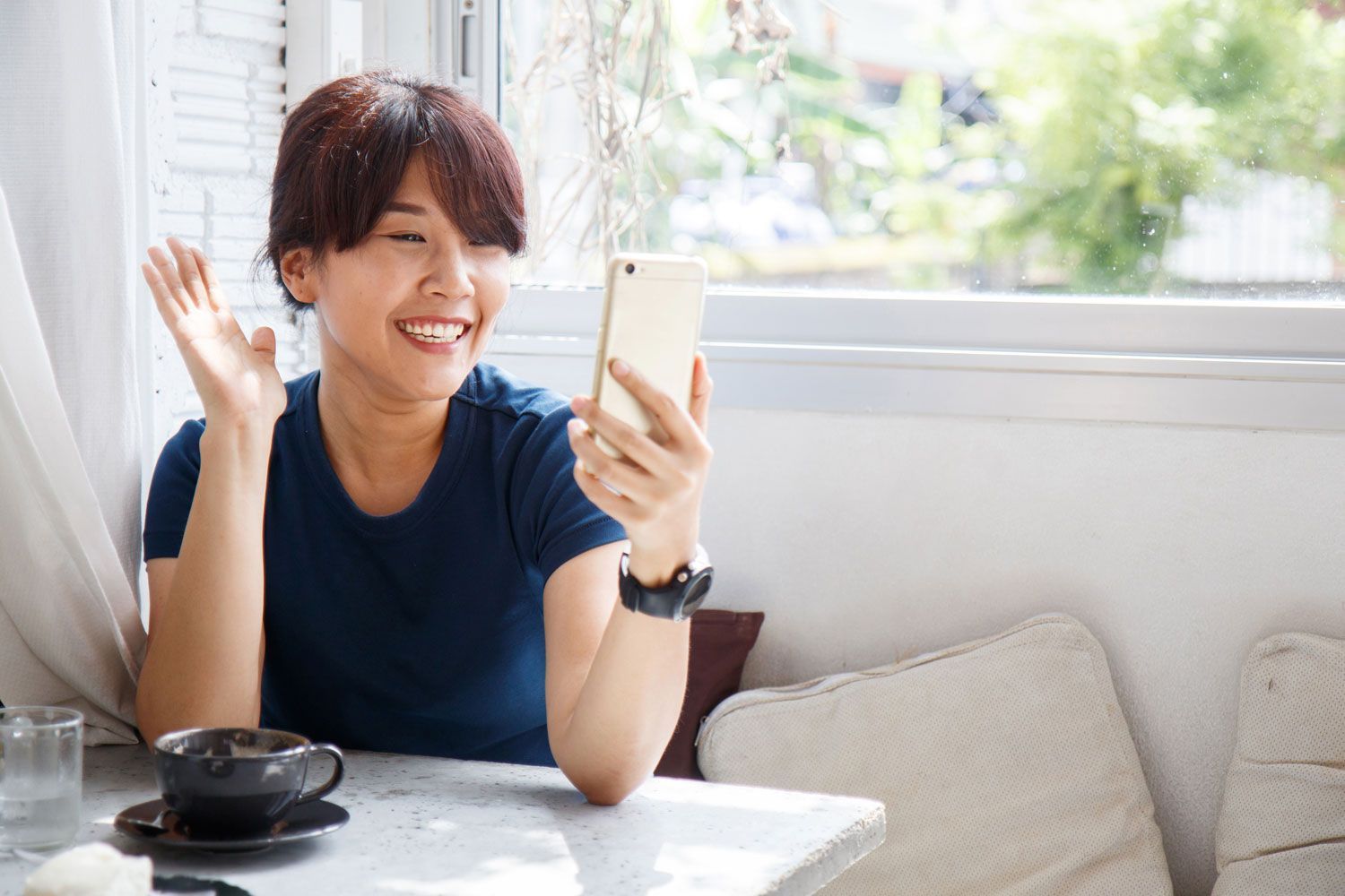 Woman in blue shirt waves during a video call, holding a phone in a cafe near a window.