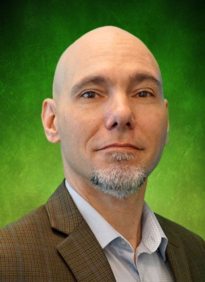 Bald man with a goatee wearing a suit jacket, in front of a green backdrop.