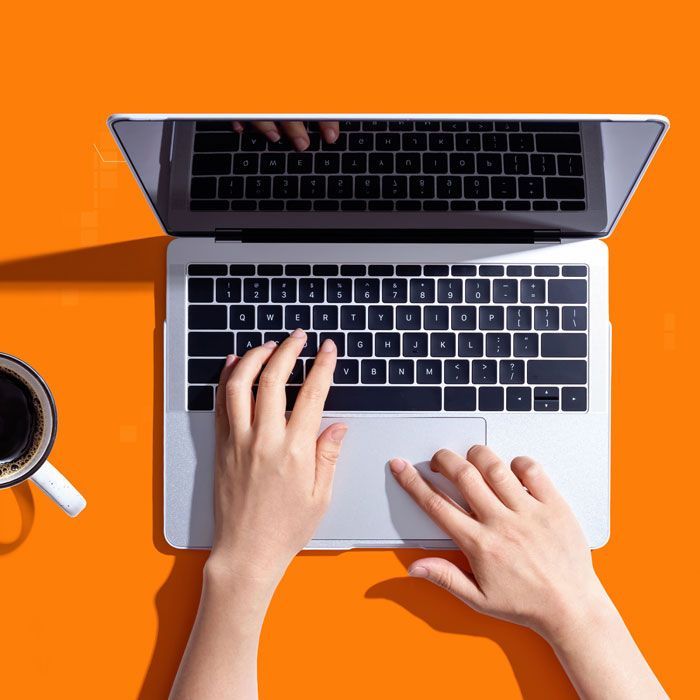 Hands typing on a laptop with a coffee cup on an orange surface.