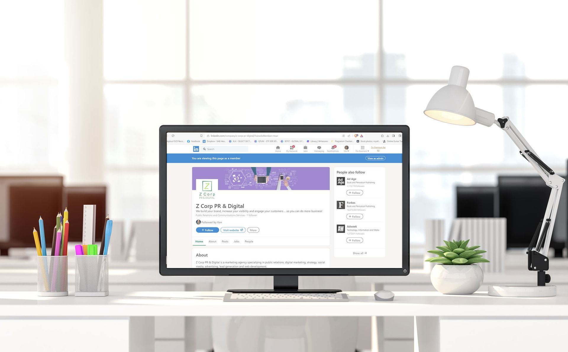 Computer screen displaying a social media profile on a bright office desk with a lamp, pens, and a plant.