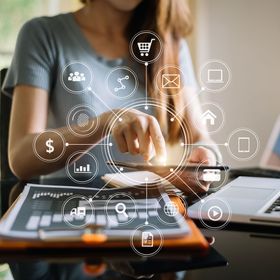 Woman uses tablet at a desk, surrounded by digital marketing icons. Laptop and graph on the desk.