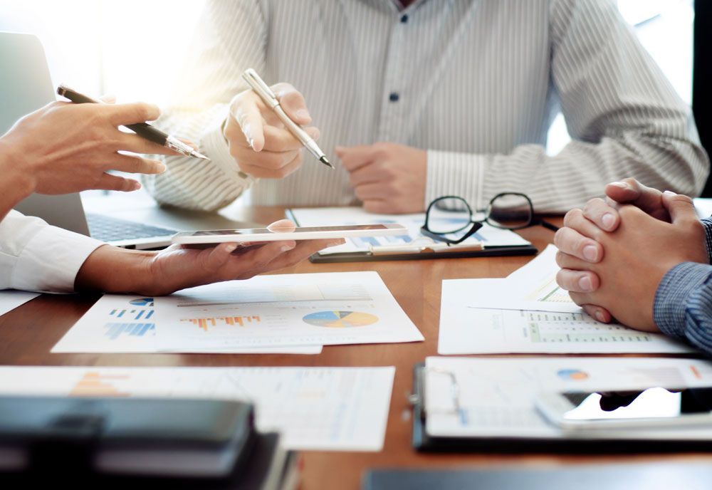 People at a table, discussing documents and pointing. Pens and glasses are visible.