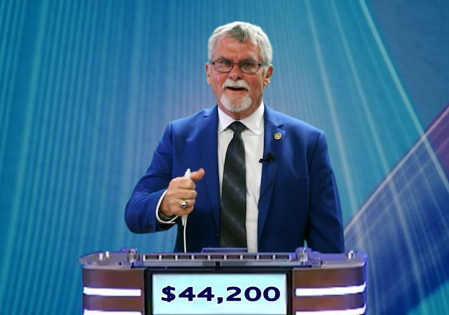Man in blue suit at podium, holding pen. Score displayed: $44,200. Blue abstract background.