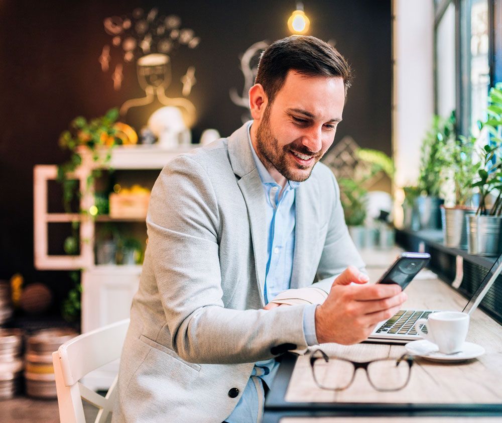 Man smiling while looking at his phone, sitting at a table with a laptop, coffee, and glasses.