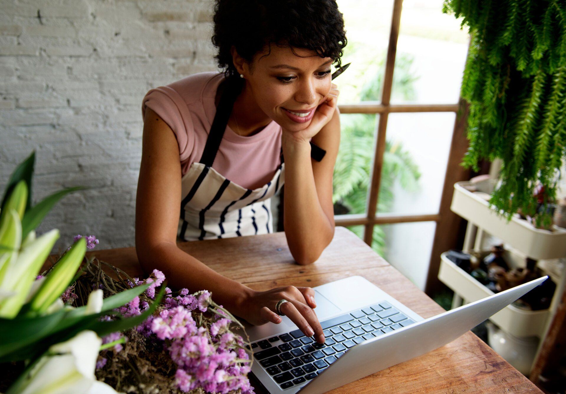 Woman in apron smiling, using laptop at a table with flowers.