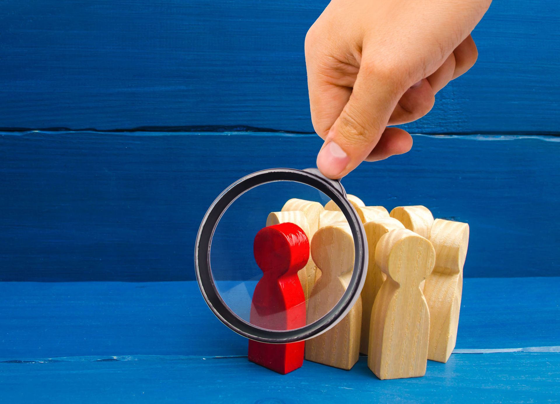 Hand holding a magnifying glass over a red figure in a group of tan figures on a blue surface.